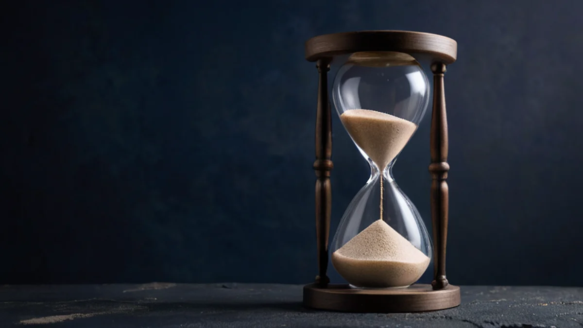 An hourglass with pale sand standing on dark stone in cool light.