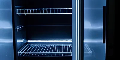 An open refrigerator door with empty glass shelves glowing in cool blue light in a dark room.